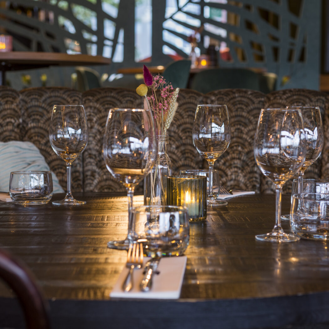 Sfeervolle gedekte houten restauranttafel met wijnglazen, bestek, vaasje droogbloemen en een brandend theelichtje.