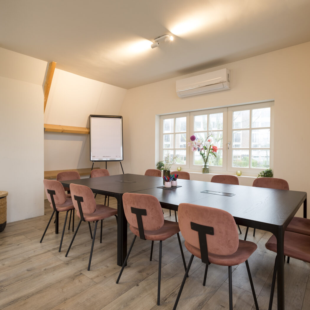 Moderne, lichte vergaderzaal met een lange zwarte tafel, comfortabele oudroze stoelen en whiteboard.