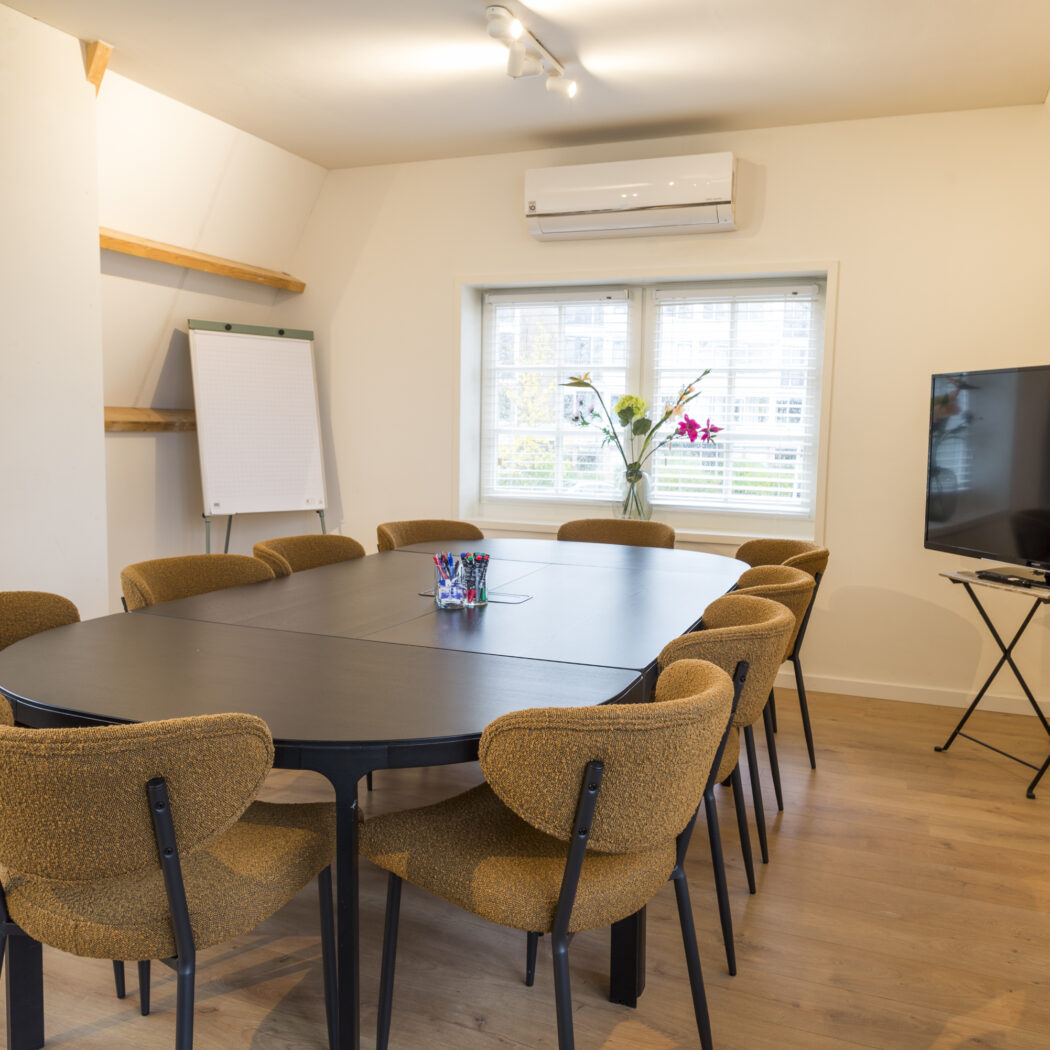 Ruime, moderne vergaderruimte met ovale zwarte tafel, mosterdgele stoelen, whiteboard en tv.