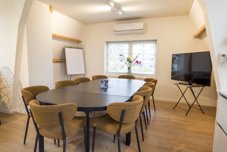 Ruime, moderne vergaderruimte met ovale zwarte tafel, mosterdgele stoelen, whiteboard en tv.