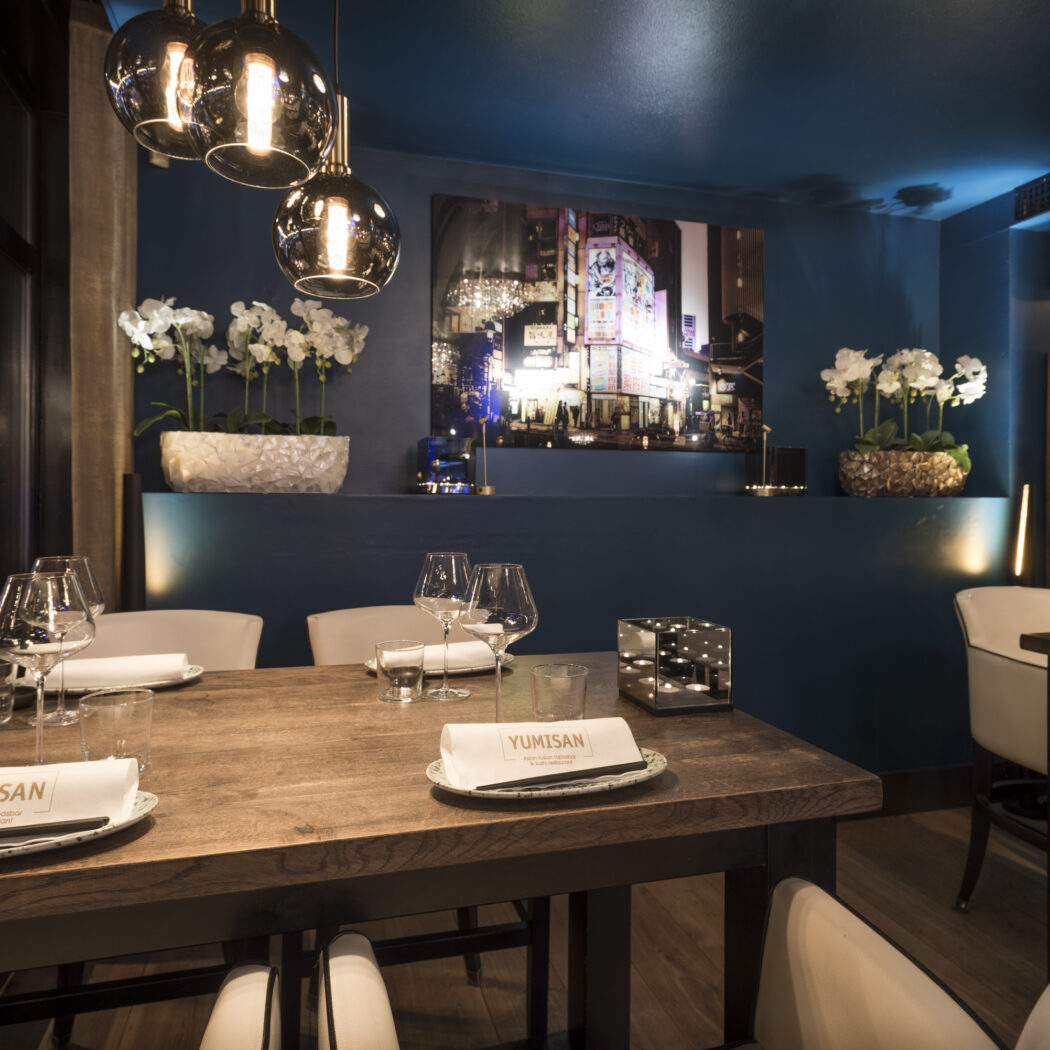 Gedekte houten restauranttafel met YUMISAN servetten, moderne hanglampen, blauwe muren en witte orchideeën.
