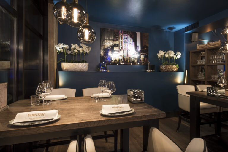 Gedekte houten restauranttafel met YUMISAN servetten, moderne hanglampen, blauwe muren en witte orchideeën.