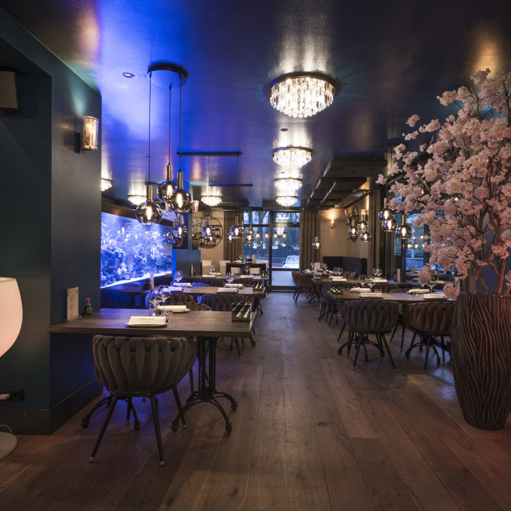 Luxe restaurantinterieur met blauw verlicht aquarium, roze bloesembomen, lichtgevende wijnglazen en moderne kroonluchters.