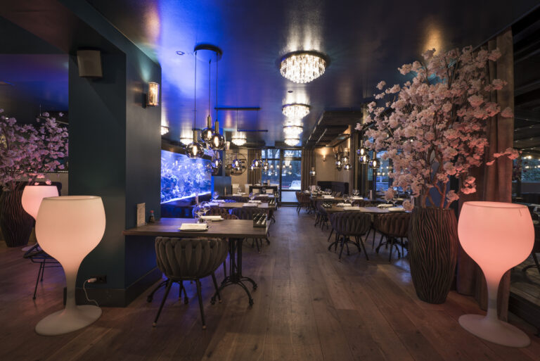 Luxe restaurantinterieur met blauw verlicht aquarium, roze bloesembomen, lichtgevende wijnglazen en moderne kroonluchters.