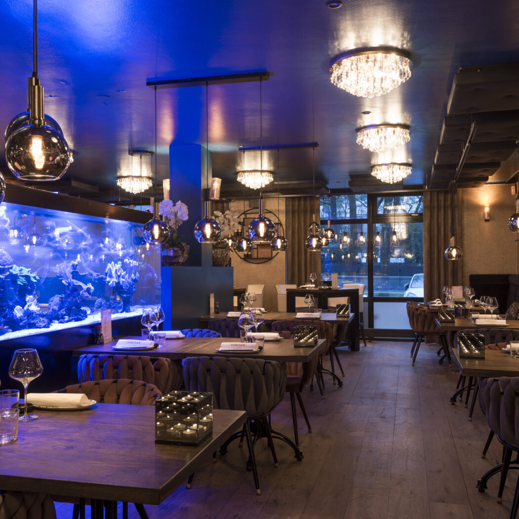 Sfeervol restaurant met groot blauw aquarium, gedekte houten tafels, diverse hanglampen en YUMISAN op servetten.