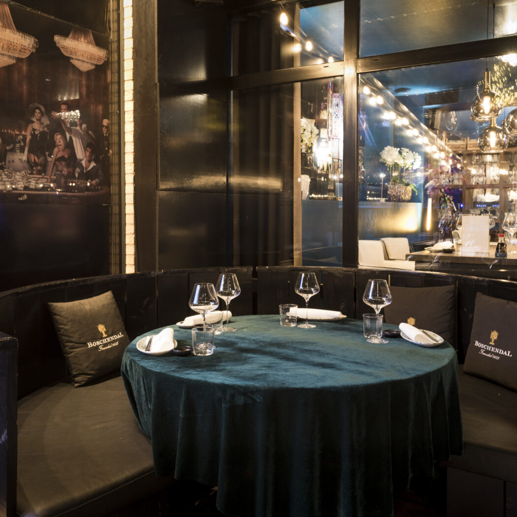 Gedekte ronde restauranttafel met donkergroen fluwelen kleed en leren zitbank, met BOSCHENDAL kussens in een sfeervol interieur.
