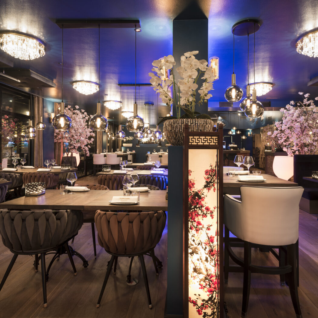 Modern restaurantinterieur met gedekte tafels, donkere muren, sfeervolle verlichting en bloemige Aziatische decoratie.