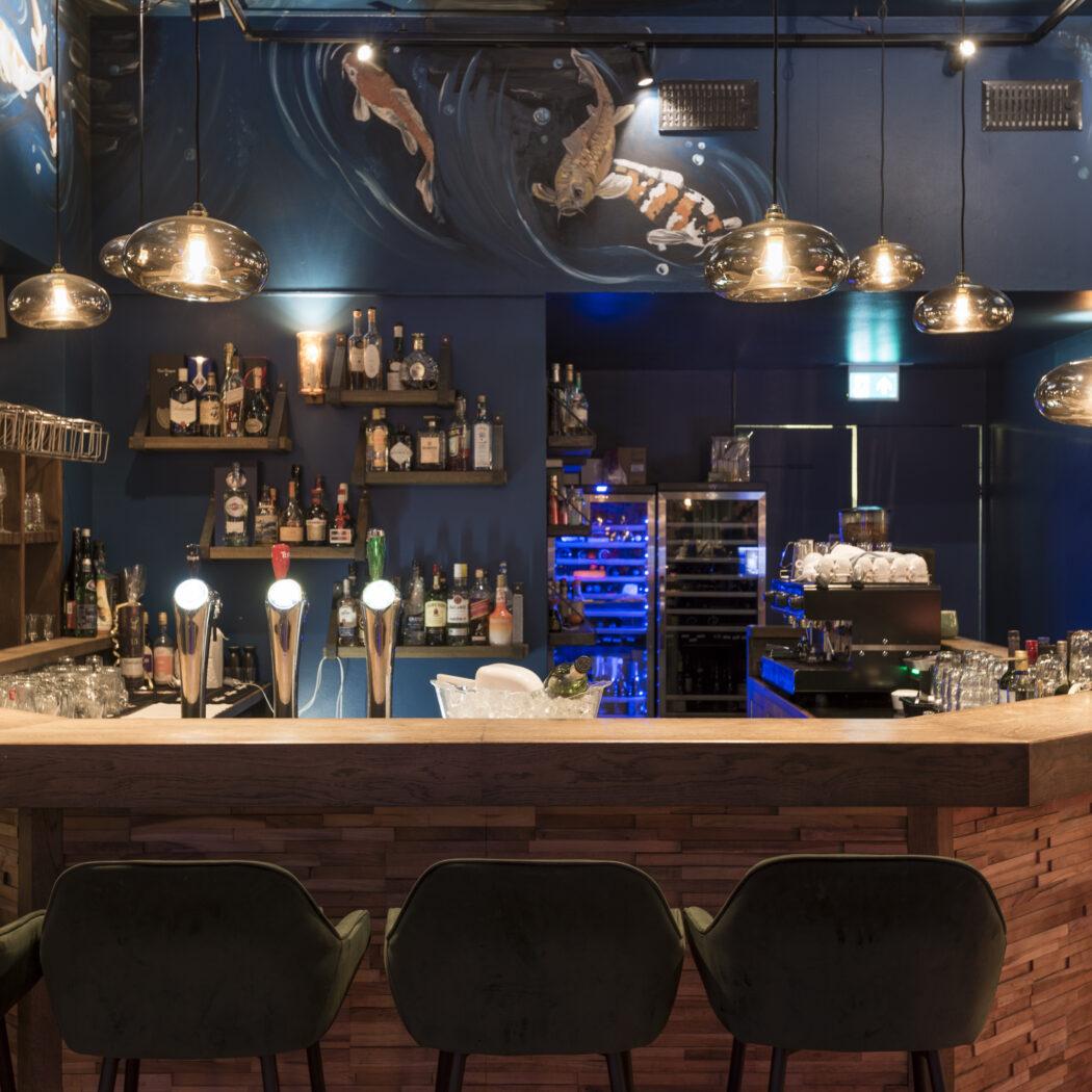 Stijlvolle houten bar met donkergroene fluwelen stoelen, bierkranen, diverse flessen en koivissen op de donkerblauwe muur.