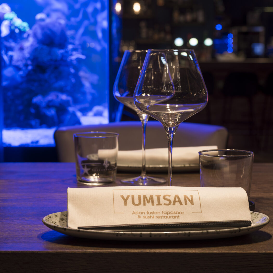 Gedekte restauranttafel met YUMISAN servet, glazen, eetstokjes en flesje sojasaus, voor een blauw verlicht aquarium.