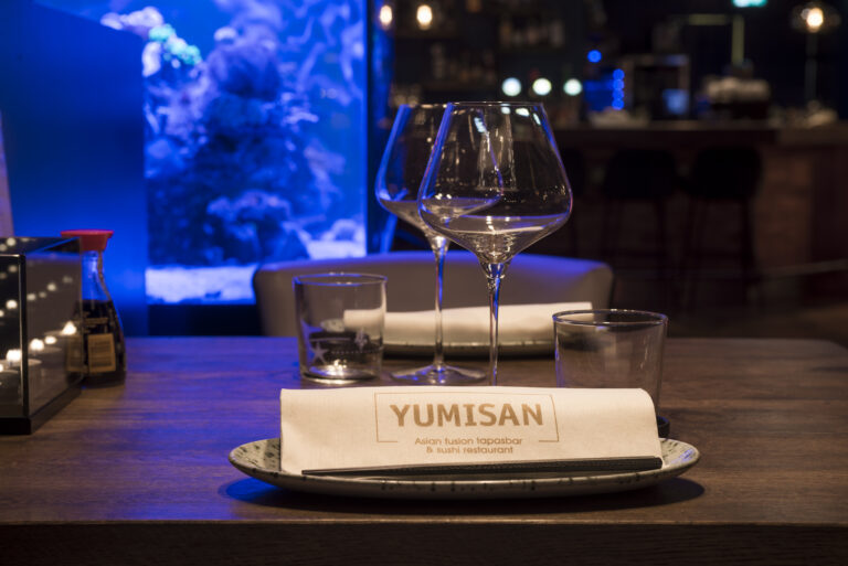 Gedekte restauranttafel met YUMISAN servet, glazen, eetstokjes en flesje sojasaus, voor een blauw verlicht aquarium.