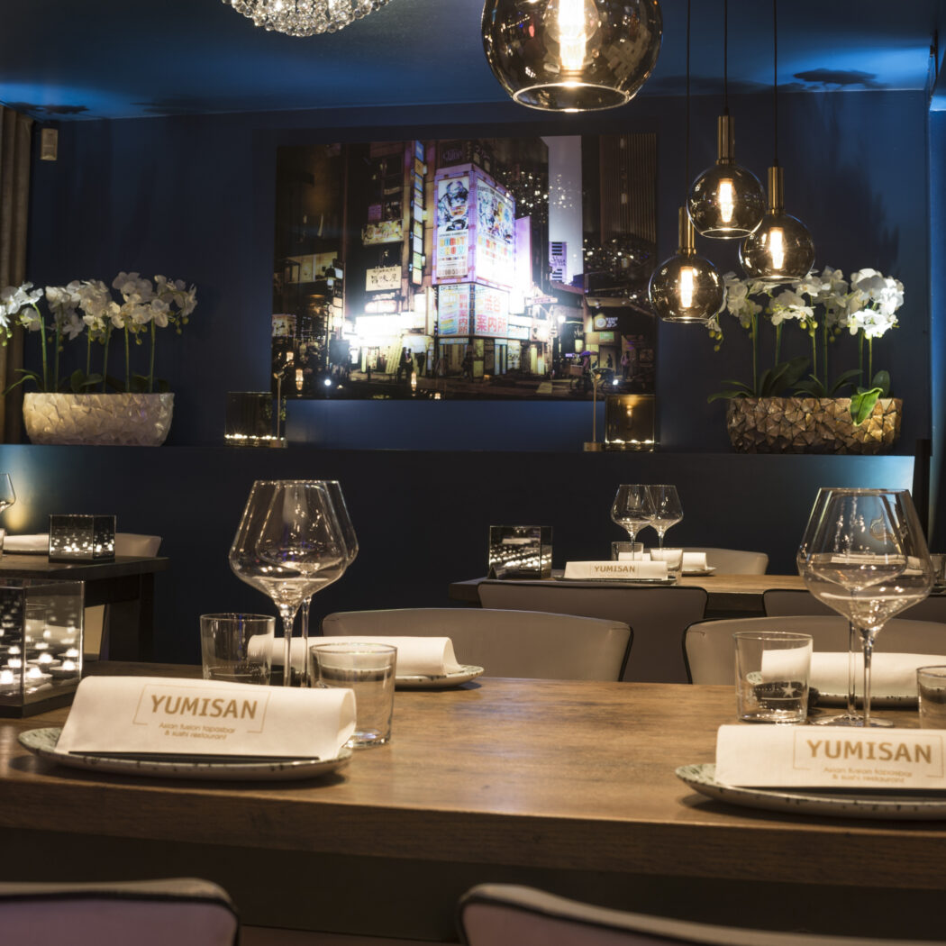 Stijlvol restaurantinterieur in donkerblauw met gedekte tafels, YUMISAN servetten en een verlichte stadsposter.