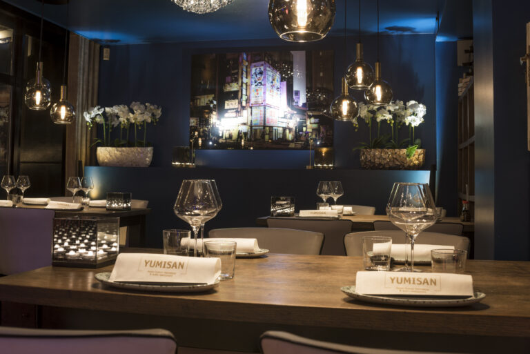 Stijlvol restaurantinterieur in donkerblauw met gedekte tafels, YUMISAN servetten en een verlichte stadsposter.