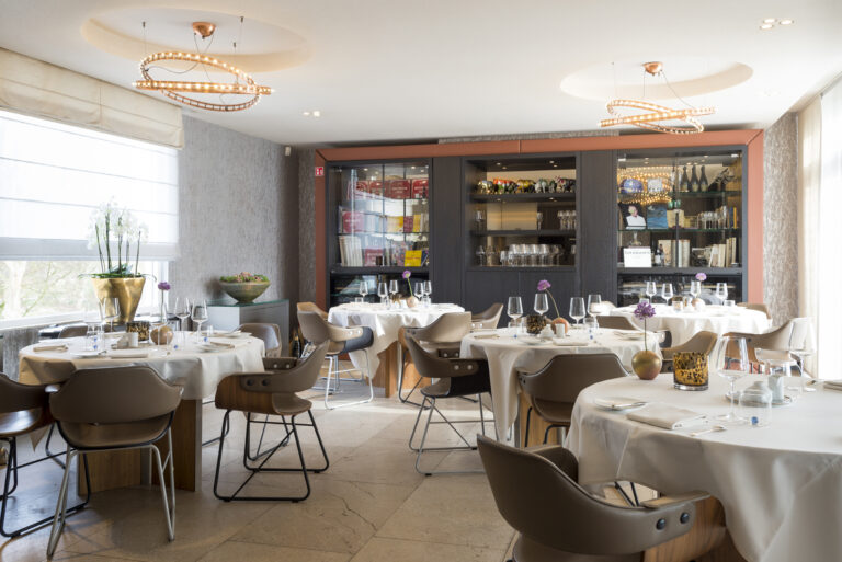 Lichte, elegante restaurant eetzaal met gedekte ronde tafels, moderne stoelen en een vitrinekast met boeken en Grahams label.