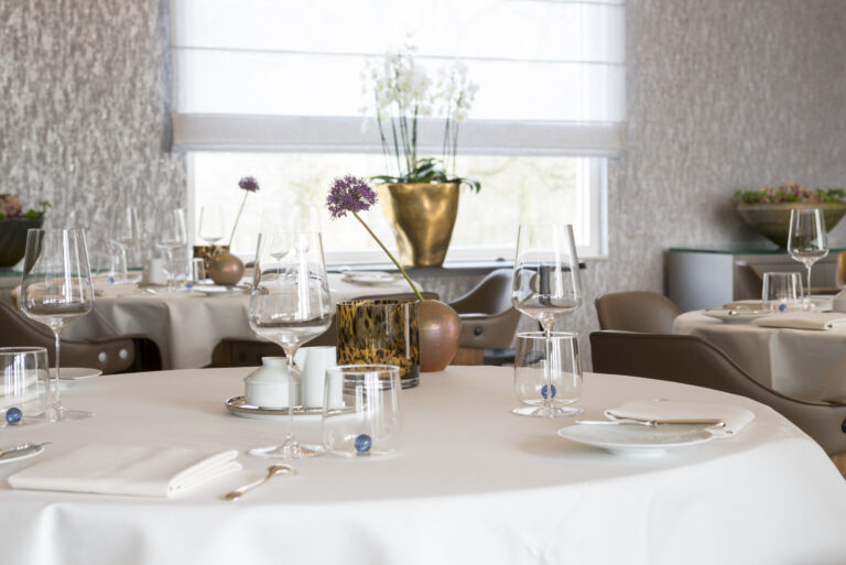 Elegant gedekte restauranttafels met witte linnen, wijnglazen, paarse allium bloemen en een raam met orchideeën.