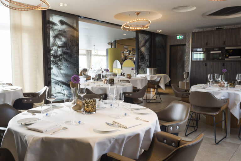 Wit gedekte tafels en bruine leren stoelen in een elegant restaurant met abstracte wandpanelen.