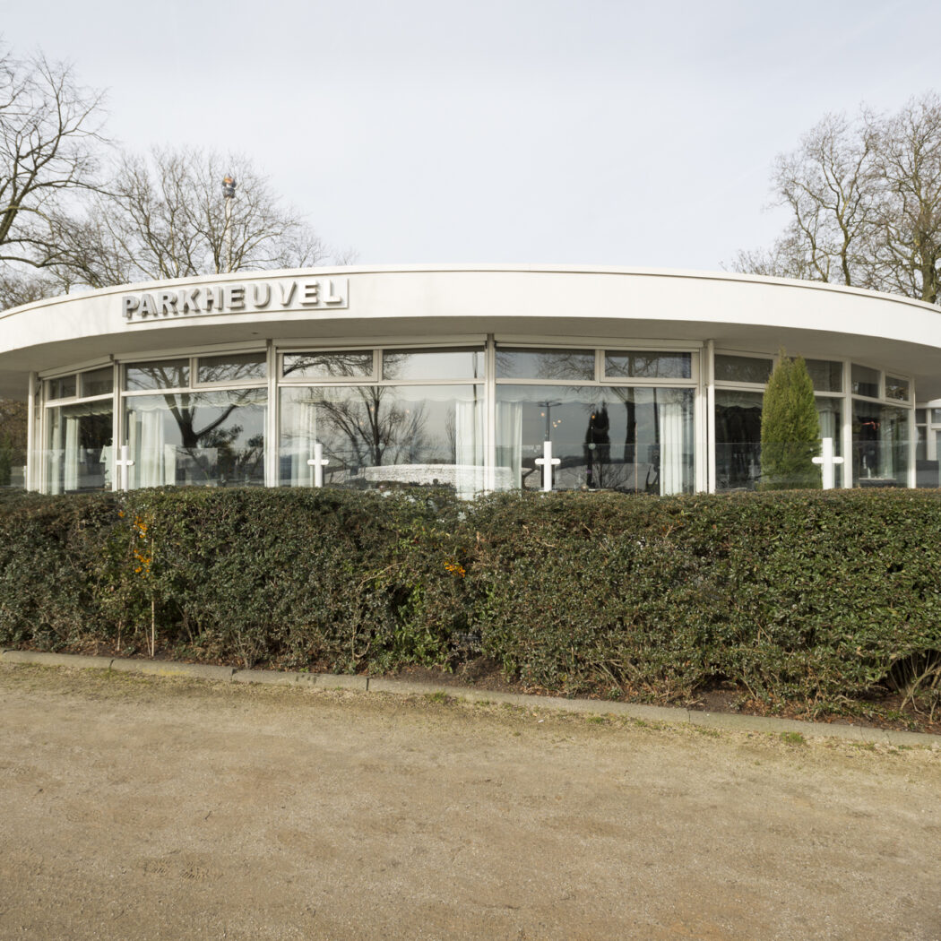 Modern wit gebouw PARKHEUVEL met rond dak en glazen gevel, omringd door kale bomen en een heg.