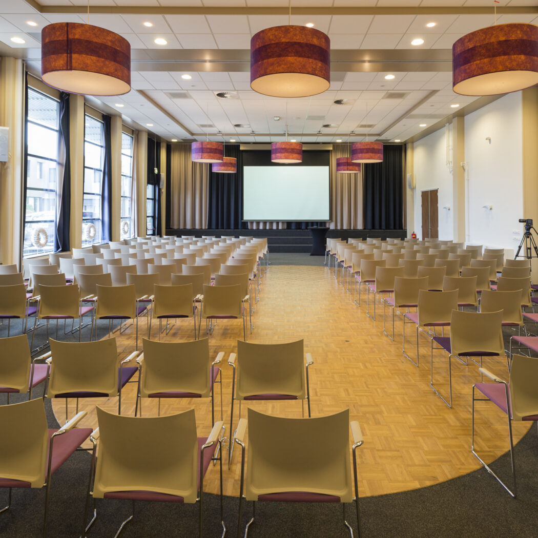 Ruime conferentiezaal met rijen beige stoelen, paarse zittingen, wit projectiescherm en gestreepte plafondlampen.