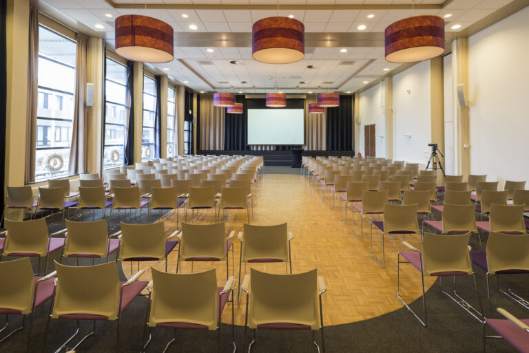 Ruime conferentiezaal met rijen beige stoelen, paarse zittingen, wit projectiescherm en gestreepte plafondlampen.