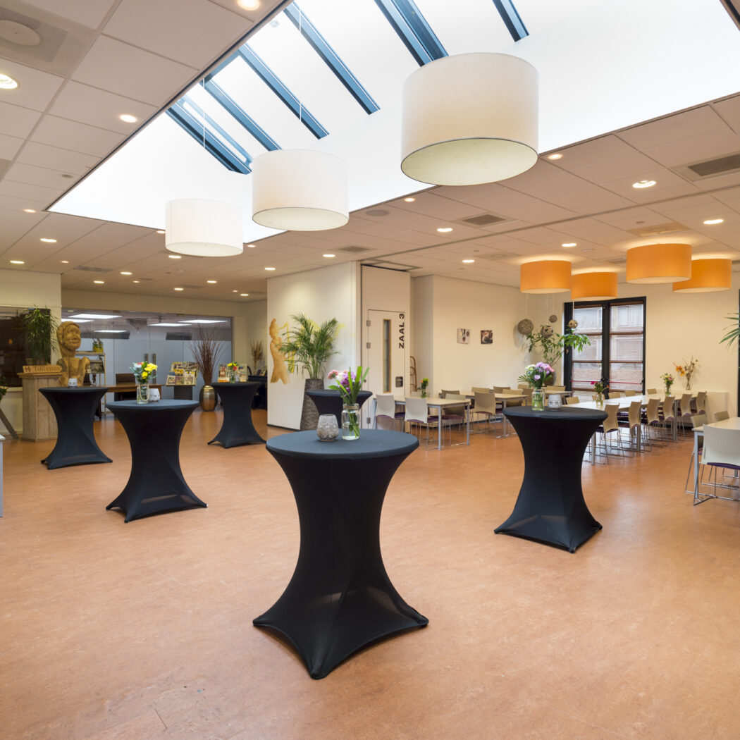 Lichte evenementenruimte met zwarte statafels, bloemstukken en tafels met stoelen onder een dakraam.