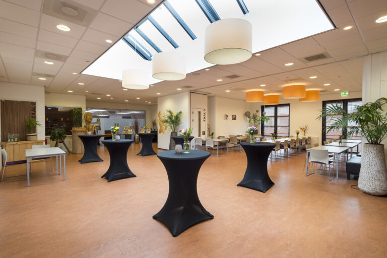 Lichte evenementenruimte met zwarte statafels, bloemstukken en tafels met stoelen onder een dakraam.