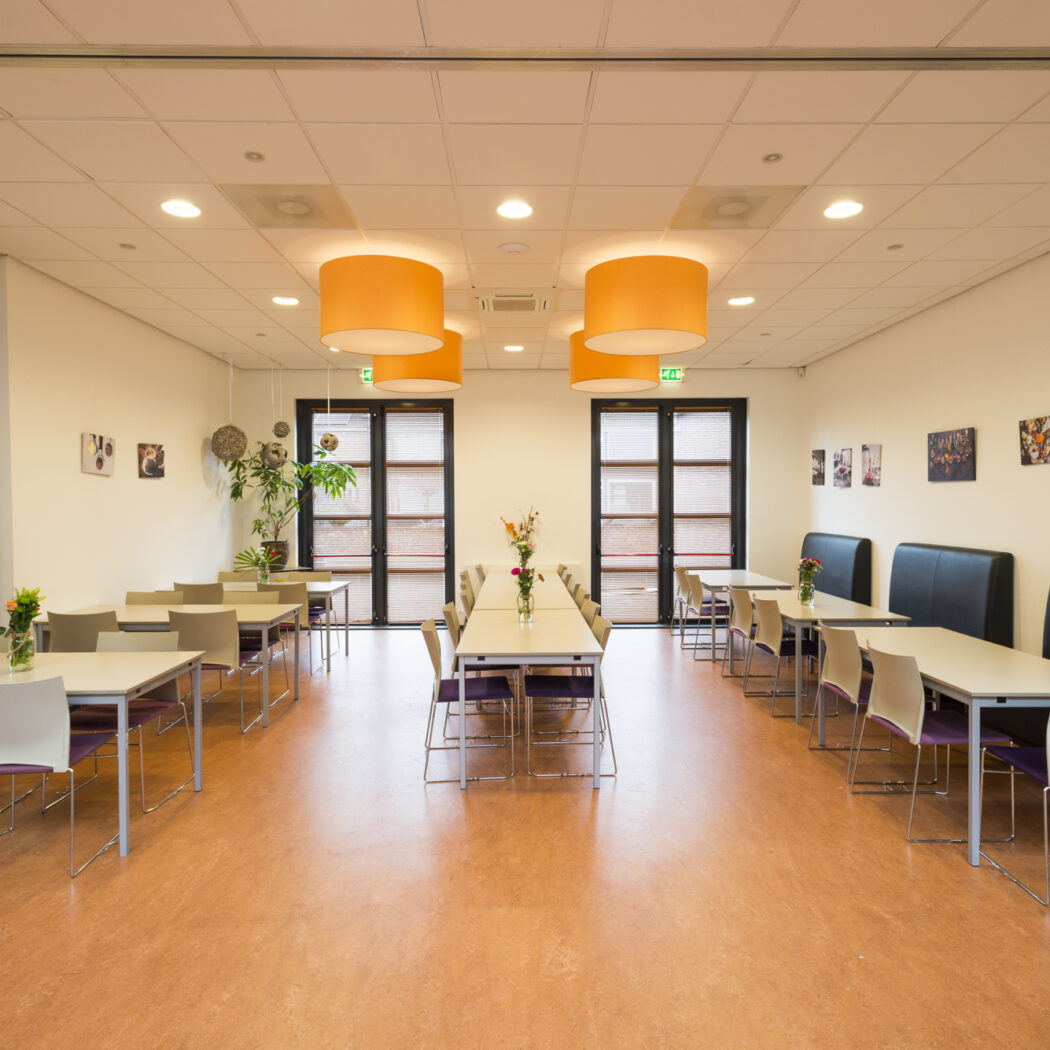 Ruime eetzaal met tafels, stoelen, zwarte banken, oranje hanglampen en “ZAAL 3” op de witte muur.