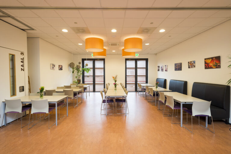 Ruime eetzaal met tafels, stoelen, zwarte banken, oranje hanglampen en “ZAAL 3” op de witte muur.