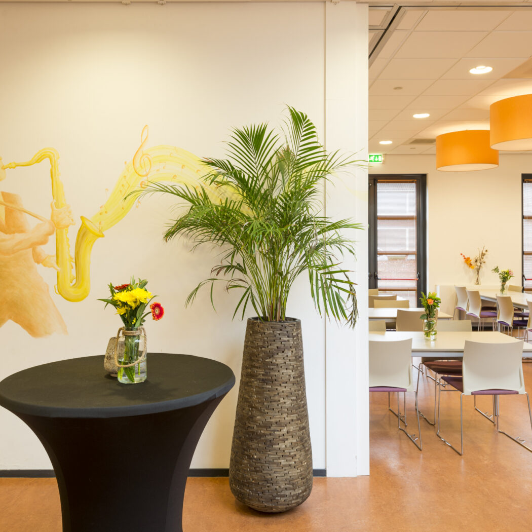 Ruime lichte evenementenruimte met een muurschildering van een saxofonist, een zwarte statafel met bloemen en lange eettafels.