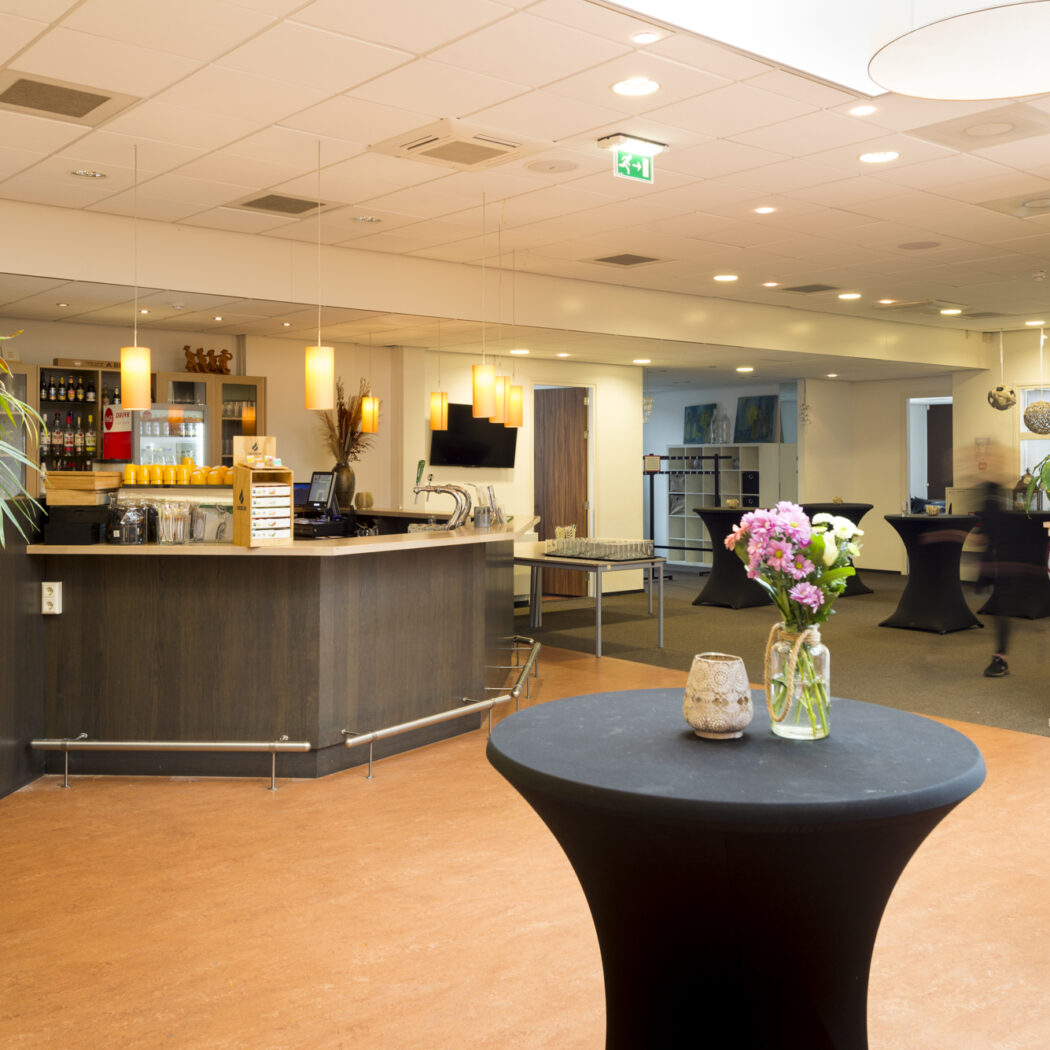 Sfeervolle receptieruimte met een houten bar, diverse zwarte statafels met bloemdecoratie en een bewegend figuur.