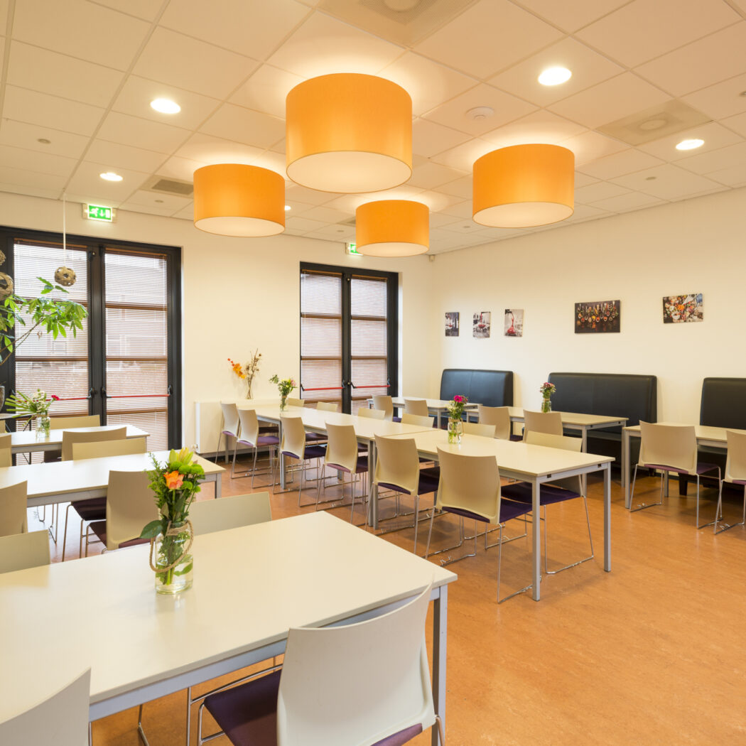Grote, lichte eetruimte met lange tafels, zwarte banken en oranje plafondlampen.