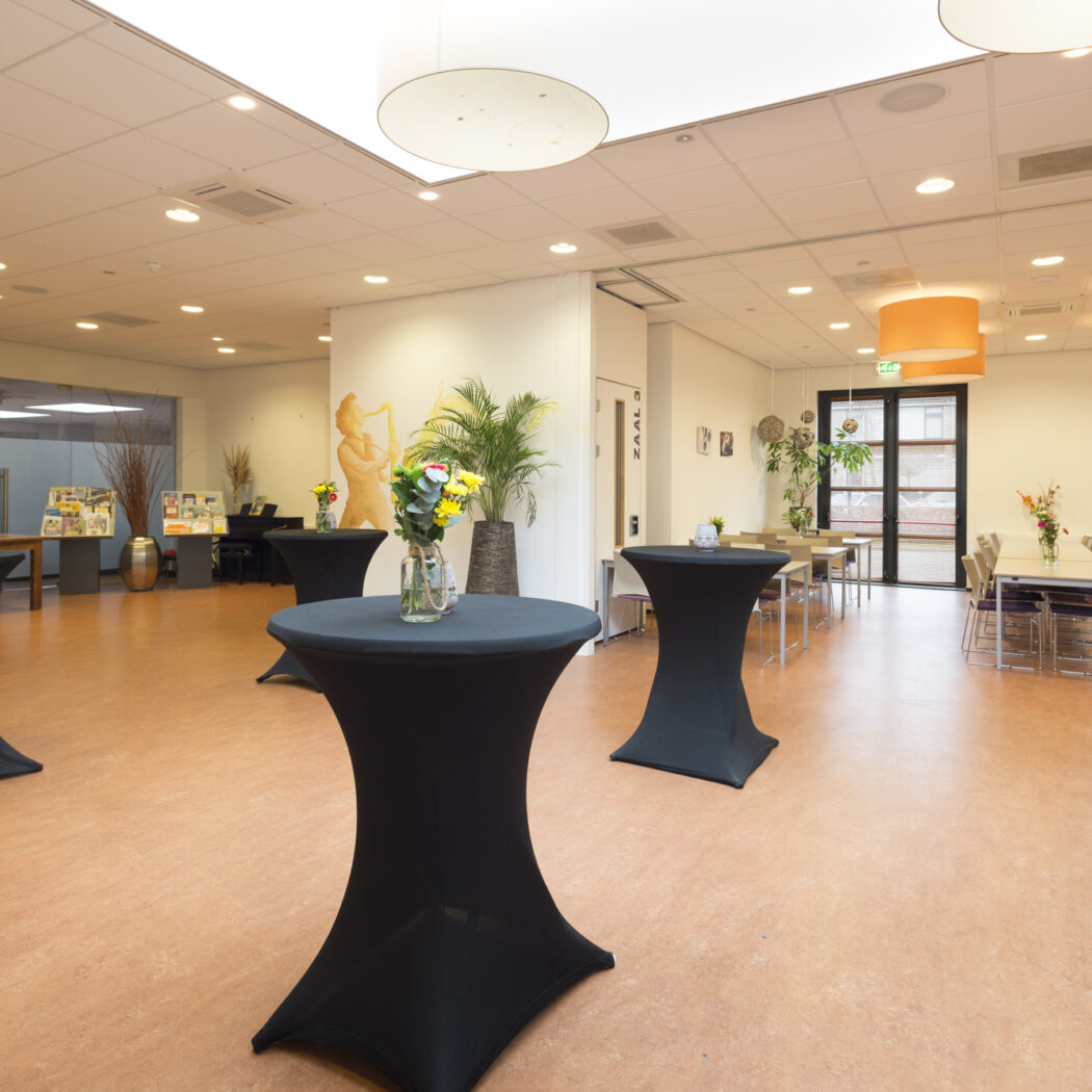 Ruime evenementenlocatie met zwarte statafels, eettafels, bloemdecoraties en oranje lampen. Borden Toiletten, ZAAL 3.
