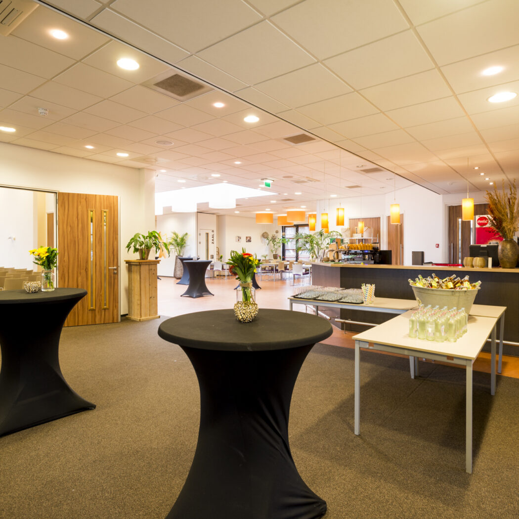 Lichte eventruimte met een bar vol dranken, zwarte staantafels en een open vergaderzaal met stoelen.