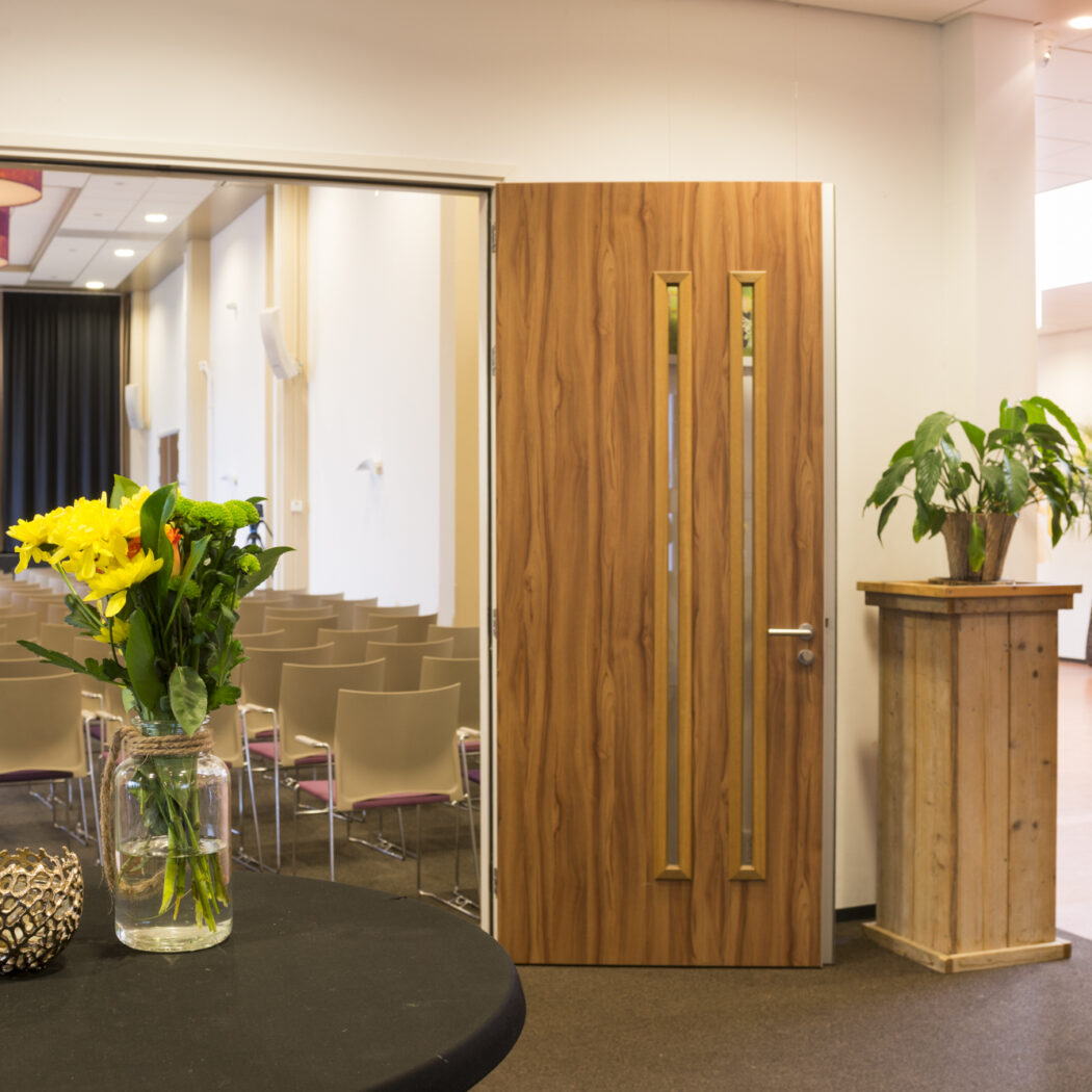 Foyer met gele bloemen op zwarte tafel en zicht op een vergaderzaal met lege stoelen; rechts entree Zaal 3.