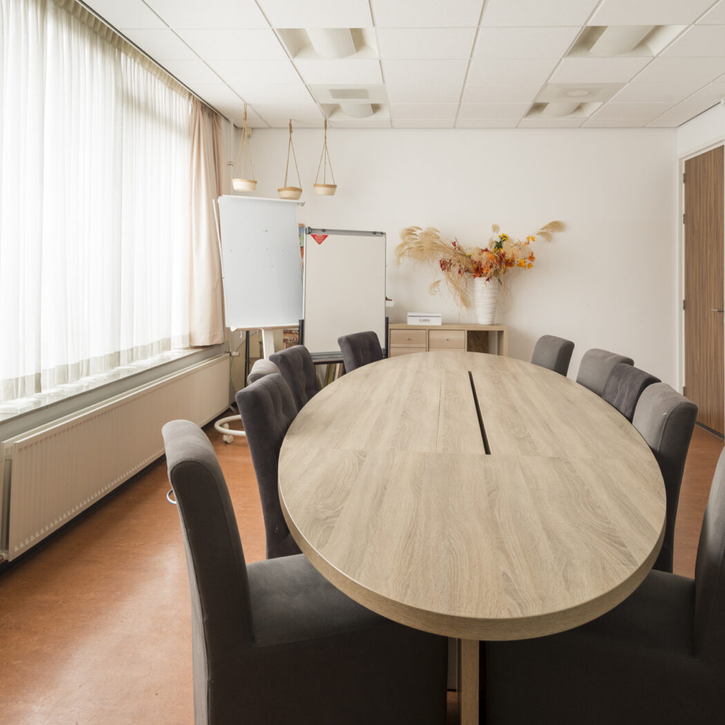 Lichte vergaderruimte met ovale lichtbruine tafel, donkergrijze stoelen, whiteboards en droogbloemdecoratie.