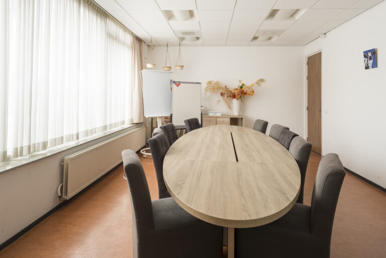 Lichte vergaderruimte met ovale lichtbruine tafel, donkergrijze stoelen, whiteboards en droogbloemdecoratie.