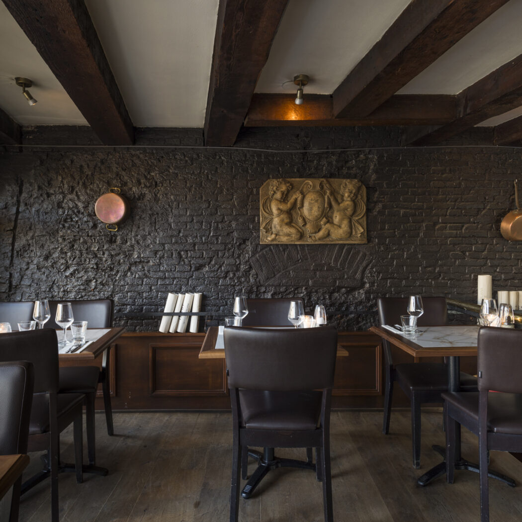 Ruim restaurant interieur met gedekte marmeren tafels, bruine lederen stoelen en een donkere bakstenen muur met houten balken.