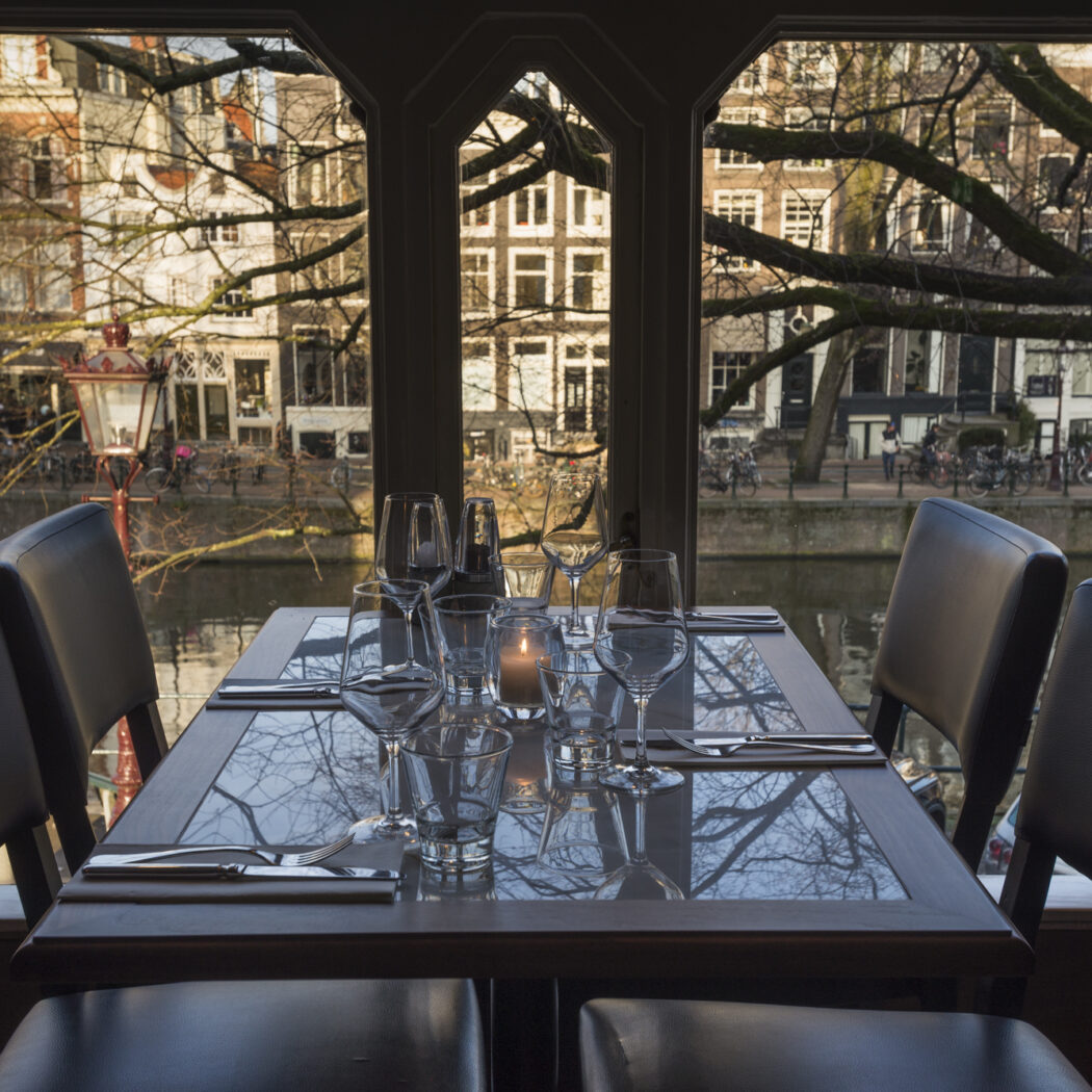 Gedekte tafel met kaarslicht en glazen in restaurant, met uitzicht op een gracht met panden, kale bomen en fietsen.