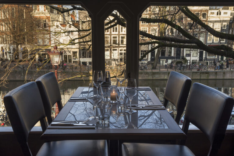Gedekte tafel met kaarslicht en glazen in restaurant, met uitzicht op een gracht met panden, kale bomen en fietsen.