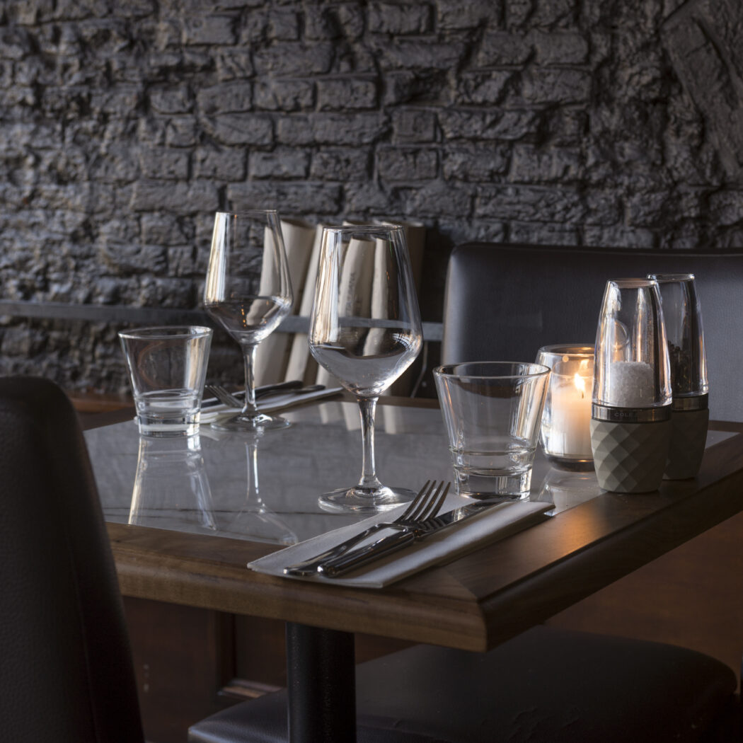 Sfeervolle gedekte restauranttafel met glazen, bestek, brandende kaars en zout- en pepermolens tegen een bakstenen muur.
