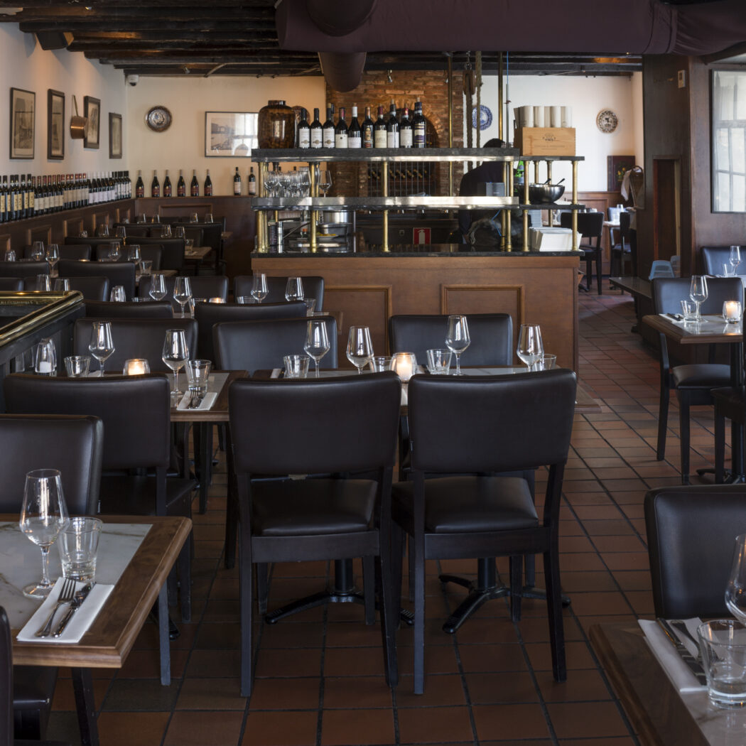 Ruim restaurantinterieur met gedekte marmeren tafels, donkere stoelen, een bar en wijnflessen op planken.