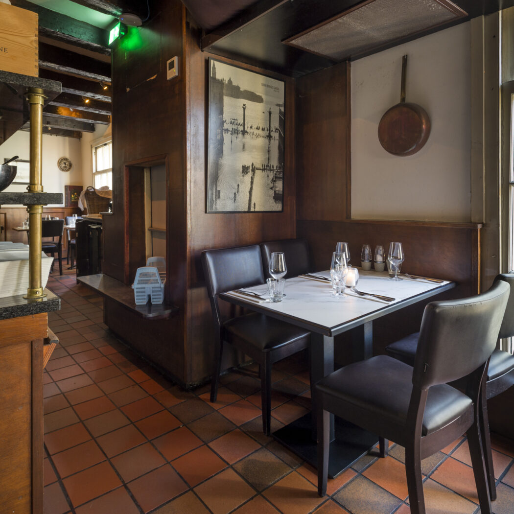 Sfeervol restaurant met gedekte tafel, donkerhout en terracotta vloer bij een groot raam.