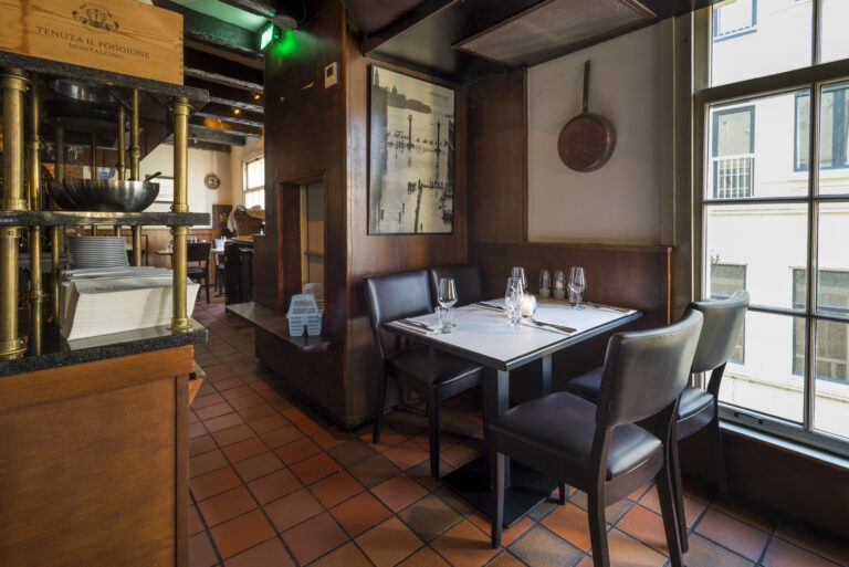 Sfeervol restaurant met gedekte tafel, donkerhout en terracotta vloer bij een groot raam.