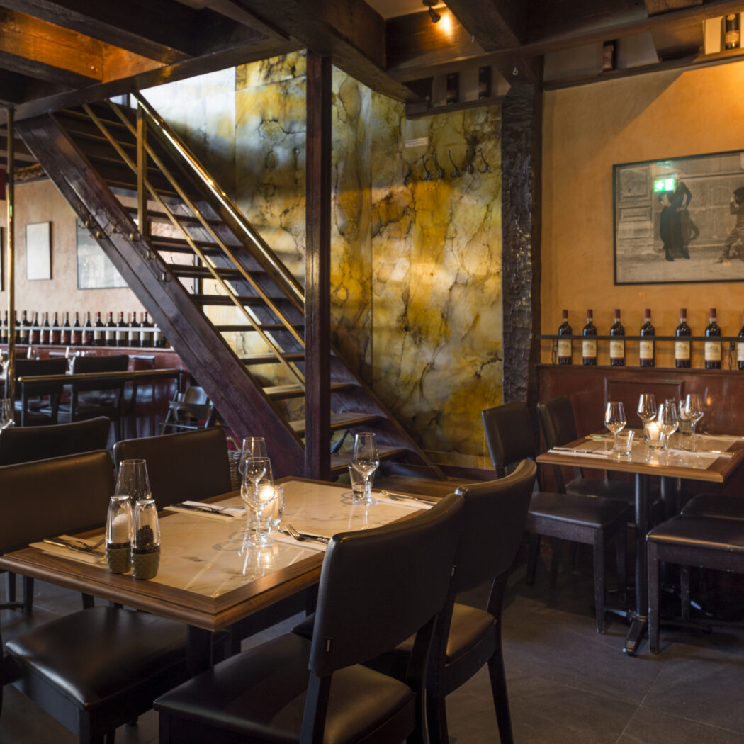 Gezellig restaurant interieur met houten trap, gedekte tafels en wijnflessen aan de muur.