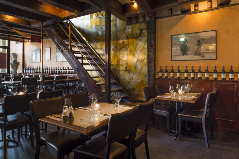 Gezellig restaurant interieur met houten trap, gedekte tafels en wijnflessen aan de muur.