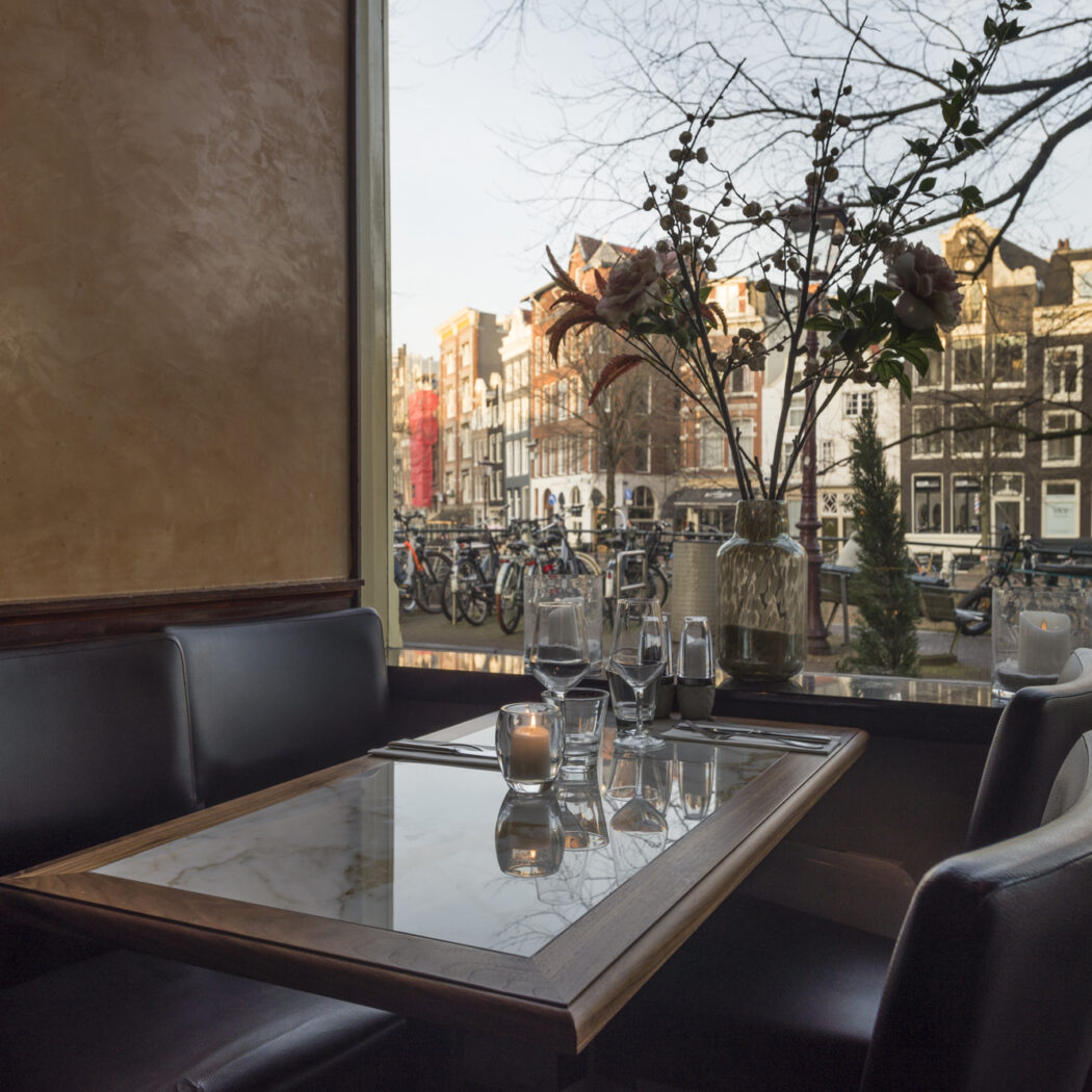 Restauranttafel met kaars en glazen aan het raam, met uitzicht op Amsterdamse grachtenpanden en fietsen.