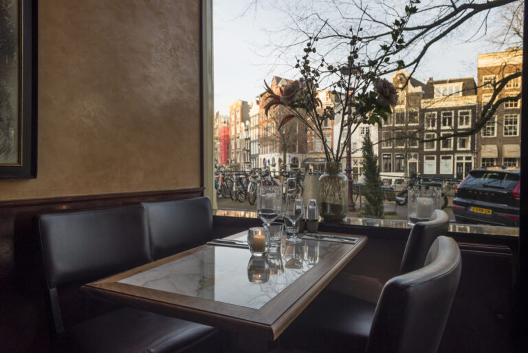 Restauranttafel met kaars en glazen aan het raam, met uitzicht op Amsterdamse grachtenpanden en fietsen.