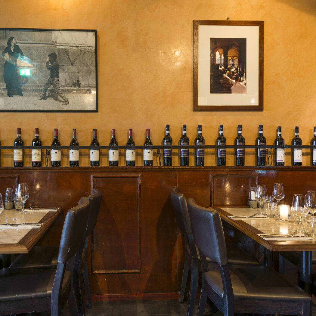 Warm restaurant interieur met okergele muren, houten lambrisering en een display van wijnflessen boven gedekte tafels.