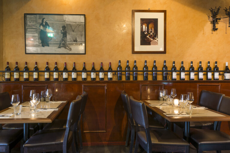 Warm restaurant interieur met okergele muren, houten lambrisering en een display van wijnflessen boven gedekte tafels.