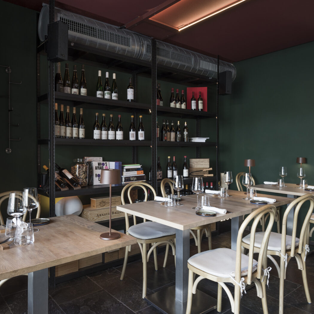 Stijlvol restaurantinterieur met gedekte houten tafels, gebogen stoelen, donkergroene muren en hoge wijnrekken.