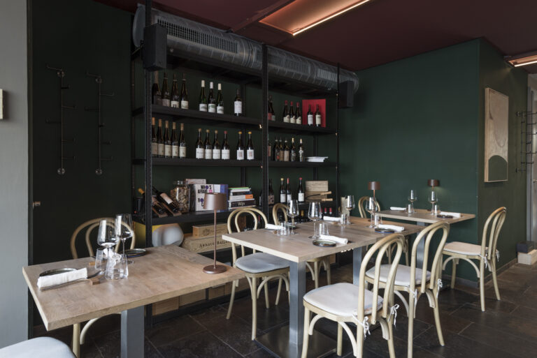 Stijlvol restaurantinterieur met gedekte houten tafels, gebogen stoelen, donkergroene muren en hoge wijnrekken.