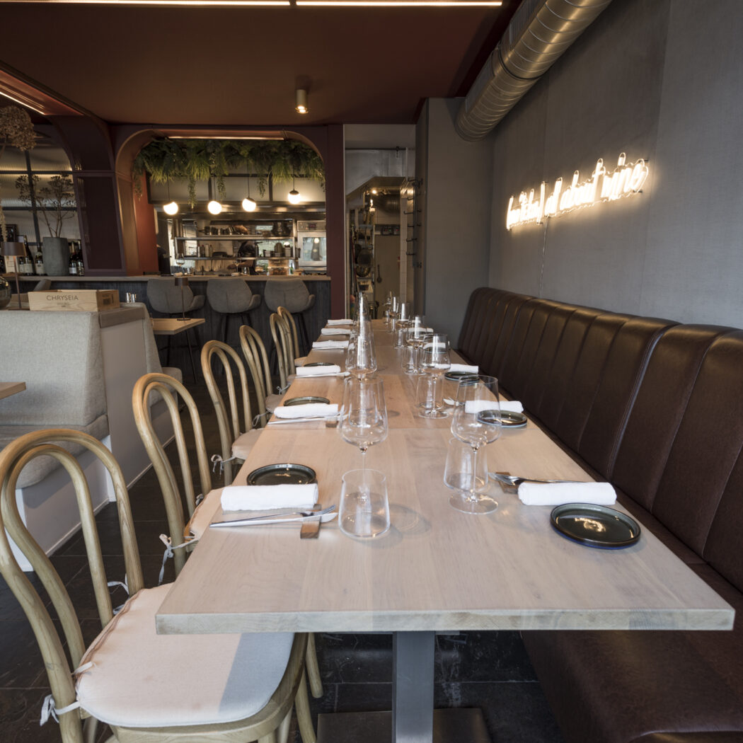 Stijlvol restaurant interieur met lange gedekte houten tafel, bruine lederen bank, rotan stoelen en neonverlichting.